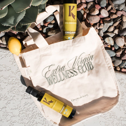 A branded tote bag with "Extra Virgin Wellness Club" embroidered on the front with pebbles and plant in the background 
